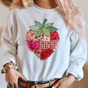 May include: A white long-sleeve sweatshirt featuring a large, central strawberry graphic. The strawberry is composed of various red and pink fabric patches with floral, heart, and gingham patterns, accented by green fabric leaves at the top.