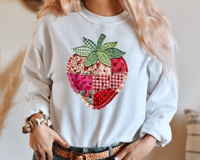 May include: A white long-sleeve sweatshirt featuring a large, central strawberry graphic. The strawberry is composed of various red and pink fabric patches with floral, heart, and gingham patterns, accented by green fabric leaves at the top.