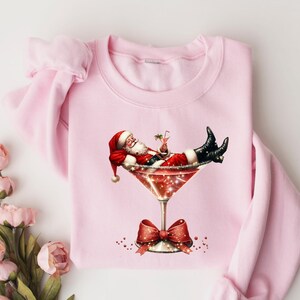 May include: Pink Christmas sweatshirt with a graphic of Santa Claus relaxing in a martini glass.