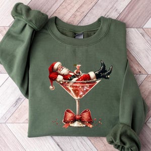 May include: Green Christmas sweatshirt with a graphic of Santa Claus relaxing in a martini glass. The glass is filled with a red liquid and has a red bow on the stem.