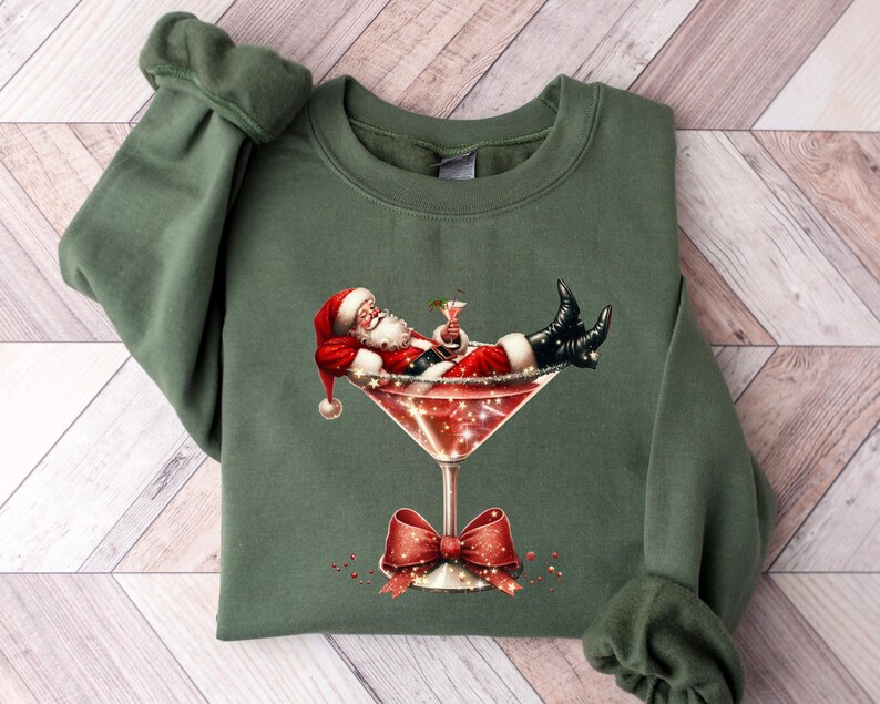May include: Green Christmas sweatshirt with a graphic of Santa Claus relaxing in a martini glass. The glass is filled with a red liquid and has a red bow on the stem.
