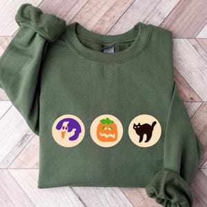 May include: A green sweatshirt with three Halloween-themed images: a ghost, a jack-o'-lantern, and a black cat. The images are in white circles on the front of the sweatshirt.