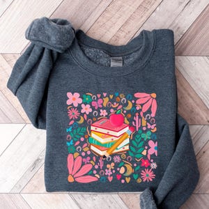 May include: A dark gray sweatshirt with a colorful graphic print of a stack of books with an apple on top. The graphic includes flowers, butterflies, and other whimsical elements.