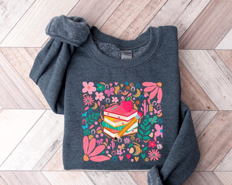 May include: A dark gray sweatshirt with a colorful graphic print of a stack of books with an apple on top. The graphic includes flowers, butterflies, and other whimsical elements.