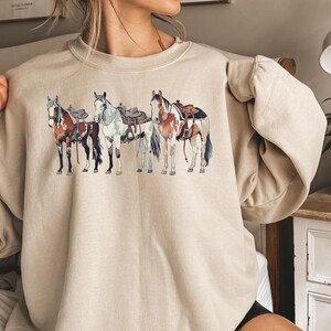 May include: A beige sweatshirt with a graphic print of four horses wearing saddles. The horses are brown, white, and black.