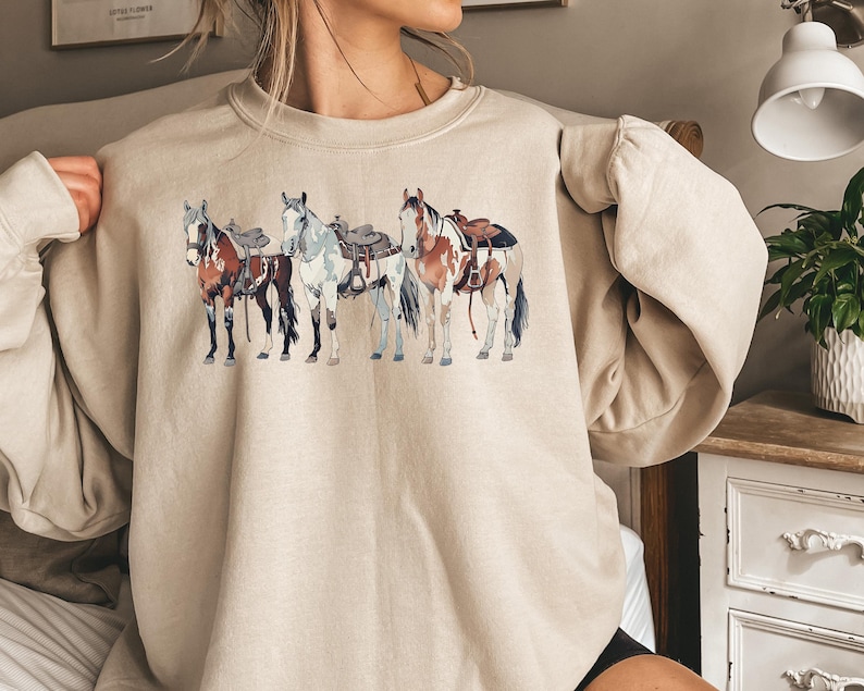 May include: A beige sweatshirt with a graphic print of four horses wearing saddles. The horses are brown, white, and black.