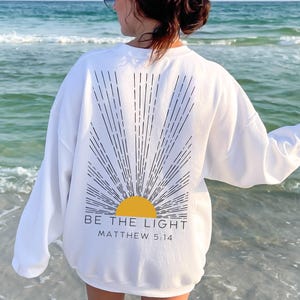 May include: White sweatshirt with a black and white sun graphic and the text "BE THE LIGHT MATTHEW 5:14".