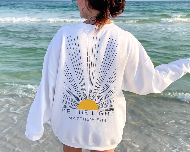May include: White sweatshirt with a black and white sun graphic and the text "BE THE LIGHT MATTHEW 5:14".
