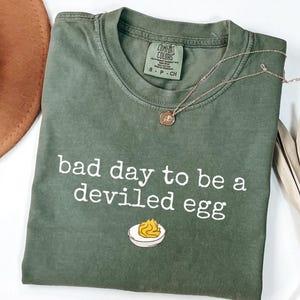 May include: Olive green t-shirt with the phrase "bad day to be a deviled egg" in white lettering. A gold necklace and a brown hat are visible. The shirt has a small graphic of a deviled egg.