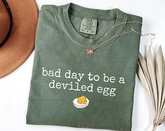 May include: Olive green t-shirt with the phrase "bad day to be a deviled egg" in white lettering. A gold necklace and a brown hat are visible. The shirt has a small graphic of a deviled egg.