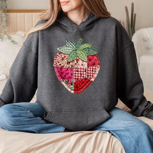May include: A dark charcoal grey hooded sweatshirt featuring a large, central strawberry graphic. The strawberry is constructed from a patchwork of various red and white fabrics, including floral, heart, and gingham patterns, with green leaves.