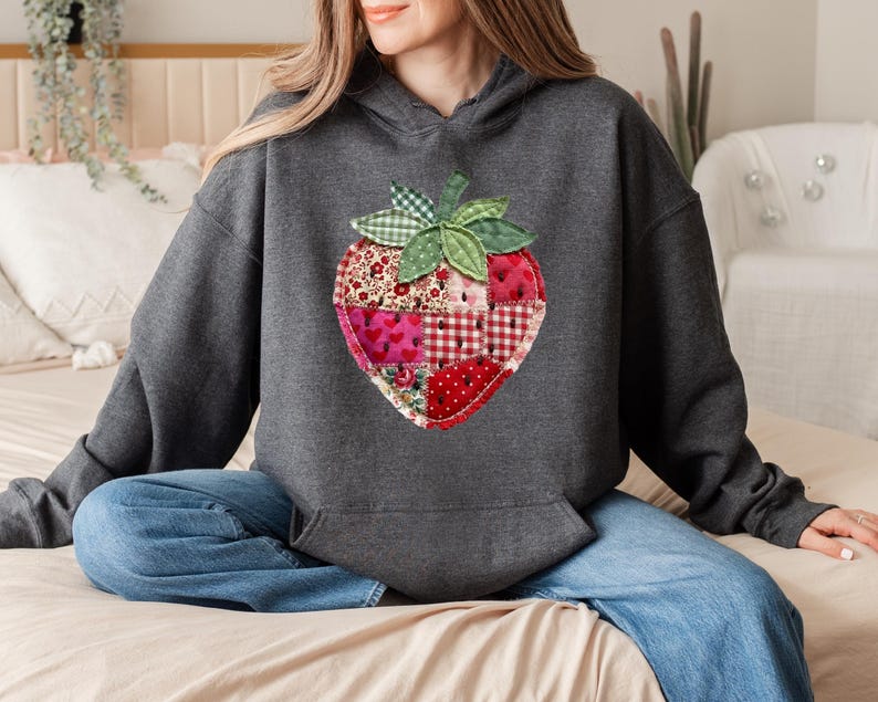 May include: A dark charcoal grey hooded sweatshirt featuring a large, central strawberry graphic. The strawberry is constructed from a patchwork of various red and white fabrics, including floral, heart, and gingham patterns, with green leaves.