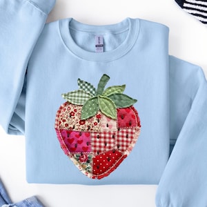 May include: A light blue crewneck sweatshirt featuring a large, central strawberry appliqu&eacute;. The strawberry is constructed from various red and white fabric patches with floral, heart, and gingham patterns, accented by green fabric leaves.