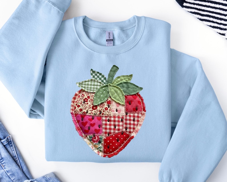 May include: A light blue crewneck sweatshirt featuring a large, central strawberry appliqu&eacute;. The strawberry is constructed from various red and white fabric patches with floral, heart, and gingham patterns, accented by green fabric leaves.