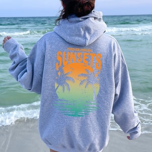 May include: Gray hooded sweatshirt with a graphic print of a sunset and palm trees. The text "FOREVER CHASING SUNSETS" is printed on the sweatshirt.