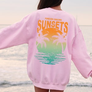 May include: Pink sweatshirt with a graphic print of palm trees and a sunset. The text "FOREVER CHASING SUNSETS" is printed on the sweatshirt.