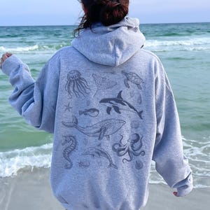May include: A gray hooded sweatshirt featuring a collection of sea creatures, including a whale, octopus, and jellyfish. The design is printed on the back of the sweatshirt. The person is standing on a beach with the ocean in the background.