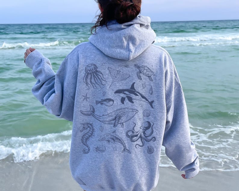 May include: A gray hooded sweatshirt featuring a collection of sea creatures, including a whale, octopus, and jellyfish. The design is printed on the back of the sweatshirt. The person is standing on a beach with the ocean in the background.