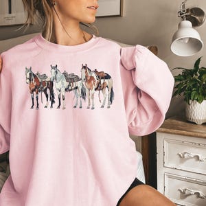 May include: A pink sweatshirt with a graphic of four horses wearing saddles. The horses are brown, white, and black.