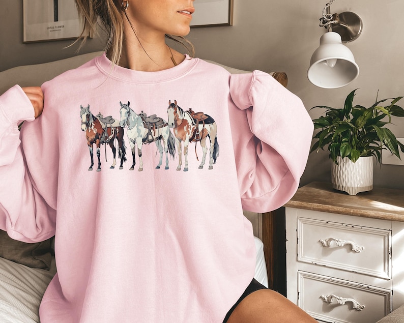 May include: A pink sweatshirt with a graphic of four horses wearing saddles. The horses are brown, white, and black.