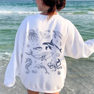 May include: White sweatshirt featuring a collection of blue sea creature illustrations, including a jellyfish, stingray, turtle, orca, whale, seahorse, dolphin, and octopus. The long-sleeved top is worn on a beach.