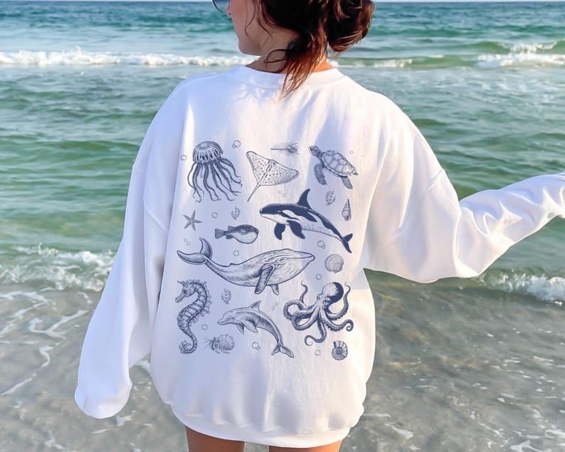 May include: White sweatshirt featuring a collection of blue sea creature illustrations, including a jellyfish, stingray, turtle, orca, whale, seahorse, dolphin, and octopus. The long-sleeved top is worn on a beach.