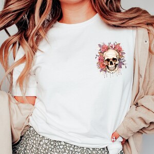 May include: White t-shirt featuring a watercolor design of a skull surrounded by pink and purple flowers. The skull has a detailed, realistic appearance with dripping watercolor effects. The design is centered on the chest of the shirt.