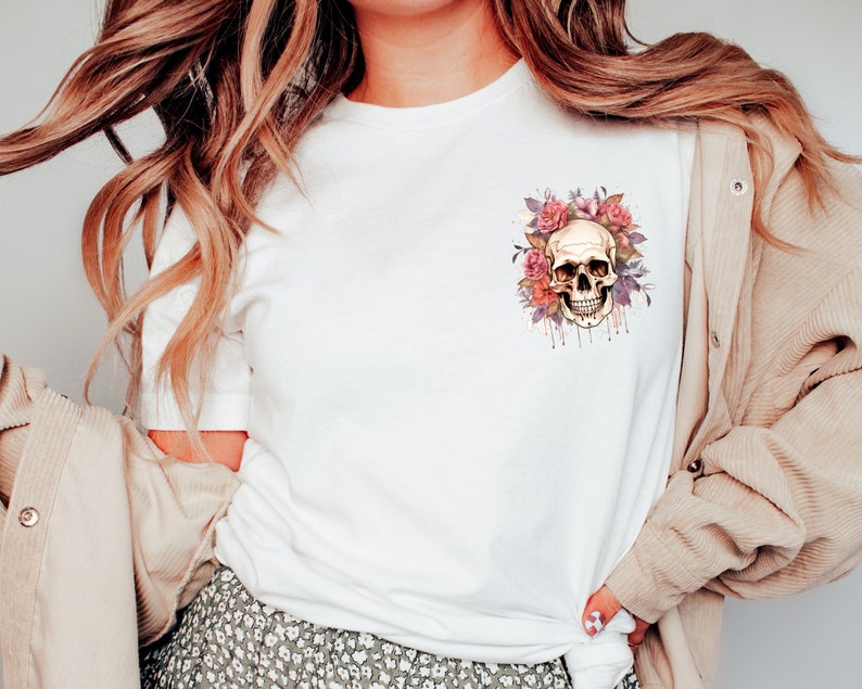 May include: White t-shirt featuring a watercolor design of a skull surrounded by pink and purple flowers. The skull has a detailed, realistic appearance with dripping watercolor effects. The design is centered on the chest of the shirt.
