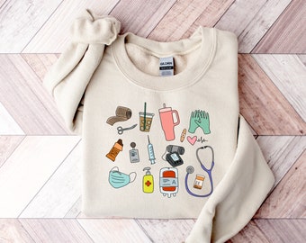 May include: A beige sweatshirt with a graphic print of medical supplies, including a stethoscope, bandages, a syringe, a blood bag, and a cup of coffee. The text "Gildan" is visible on the tag.