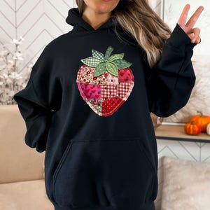 May include: A black hooded sweatshirt featuring a large, central strawberry graphic. The strawberry is designed with a patchwork-style pattern in various shades of red, pink, and white, accented by a green leaf top with a checkered texture.