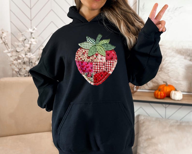 May include: A black hooded sweatshirt featuring a large, central strawberry graphic. The strawberry is designed with a patchwork-style pattern in various shades of red, pink, and white, accented by a green leaf top with a checkered texture.
