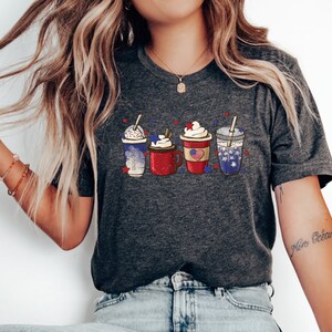 May include: Dark gray t-shirt featuring a graphic of four festive drinks in red, white, and blue, with whipped cream and star accents. The design evokes a patriotic theme, perfect for celebrating a national holiday.