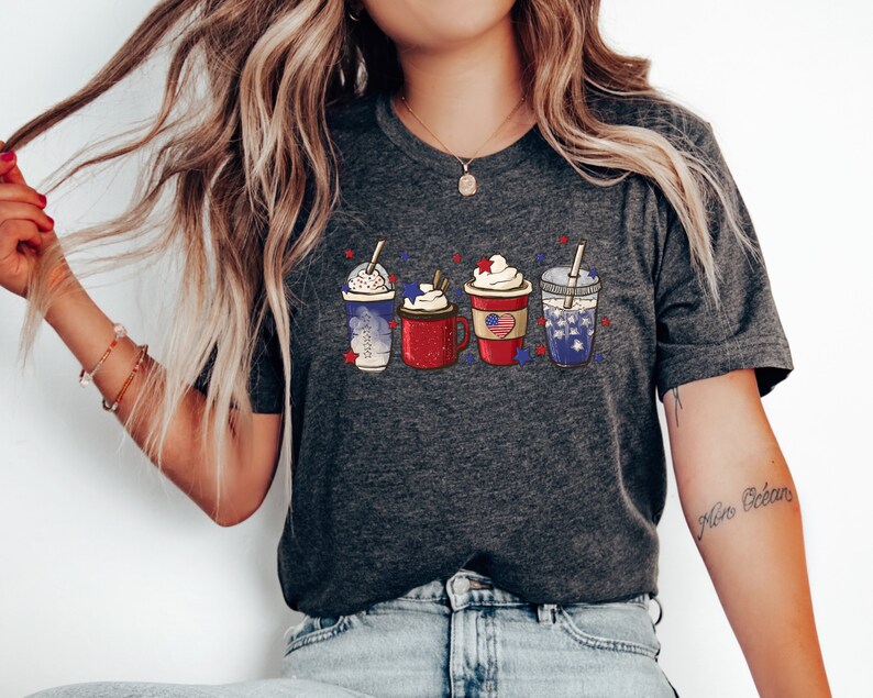 May include: Dark gray t-shirt featuring a graphic of four festive drinks in red, white, and blue, with whipped cream and star accents. The design evokes a patriotic theme, perfect for celebrating a national holiday.