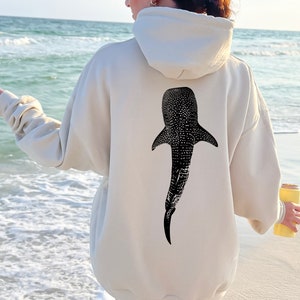 May include: Cream-colored hoodie featuring a large black and white whale shark graphic on the back. The hoodie has a hood and long sleeves. The image is taken outdoors near the ocean.