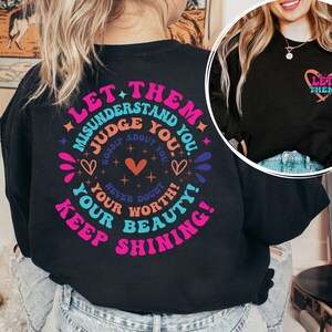 May include: Black long-sleeve shirt with a colorful graphic print that says "Let them misunderstand you. Judge about you. Gossip about you. Never doubt your worth. Your beauty! Keep shining!" and a smaller heart graphic that says "Let them" on the front.