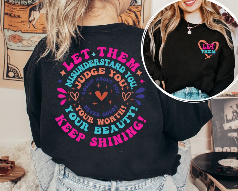 May include: Black long-sleeve shirt with a colorful graphic print that says "Let them misunderstand you. Judge about you. Gossip about you. Never doubt your worth. Your beauty! Keep shining!" and a smaller heart graphic that says "Let them" on the front.