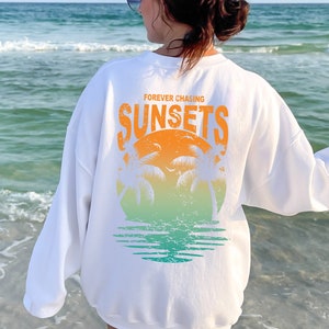 May include: White sweatshirt with a graphic print of a sunset with palm trees and the text "FOREVER CHASING SUNSETS".