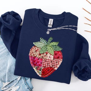 May include: A navy blue crewneck sweatshirt featuring a large, central strawberry appliqu&eacute; made from various patchwork fabric squares in shades of red, pink, and white with floral, heart, and gingham patterns, topped with green gingham leaves.