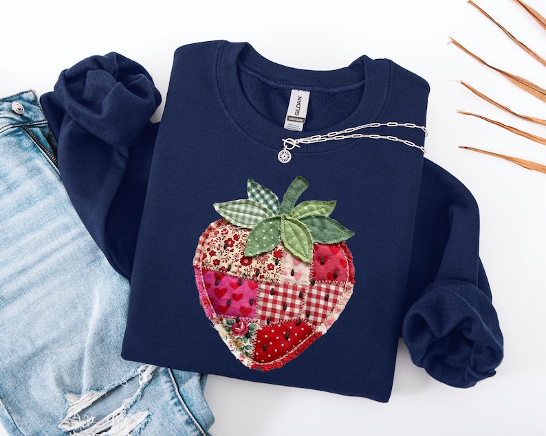 May include: A navy blue crewneck sweatshirt featuring a large, central strawberry appliqu&eacute; made from various patchwork fabric squares in shades of red, pink, and white with floral, heart, and gingham patterns, topped with green gingham leaves.