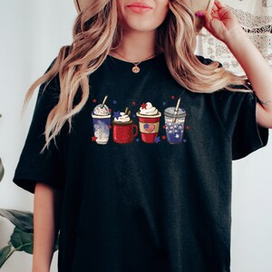 May include: Black t-shirt featuring a design of four festive drinks, each adorned with whipped cream and patriotic embellishments. The drinks include a blue and white beverage, a red mug with an American flag, and a clear cup with a straw.
