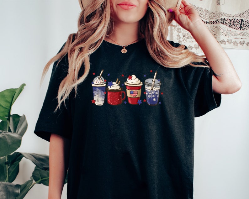 May include: Black t-shirt featuring a design of four festive drinks, each adorned with whipped cream and patriotic embellishments. The drinks include a blue and white beverage, a red mug with an American flag, and a clear cup with a straw.