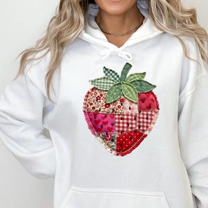 May include: A white hooded sweatshirt featuring a large, central graphic of a strawberry constructed from various patchwork fabric squares in shades of red, pink, and white, with green fabric leaves at the top of the strawberry design.