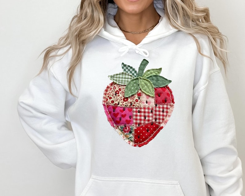 May include: A white hooded sweatshirt featuring a large, central graphic of a strawberry constructed from various patchwork fabric squares in shades of red, pink, and white, with green fabric leaves at the top of the strawberry design.