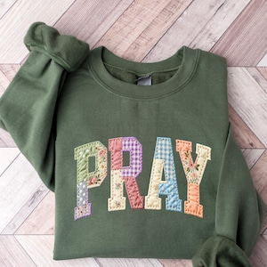 May include: An olive green sweatshirt with the word "PRAY" appliqued across the chest. The letters are made of patchwork fabric in various floral and patterned designs. The sweatshirt has long sleeves with ribbed cuffs.