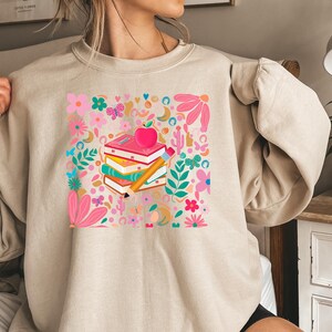May include: A beige sweatshirt with a colorful graphic print of a stack of books, a pencil, and an apple. The graphic is surrounded by flowers and other decorative elements.