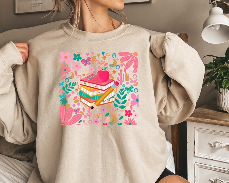 May include: A beige sweatshirt with a colorful graphic print of a stack of books, a pencil, and an apple. The graphic is surrounded by flowers and other decorative elements.