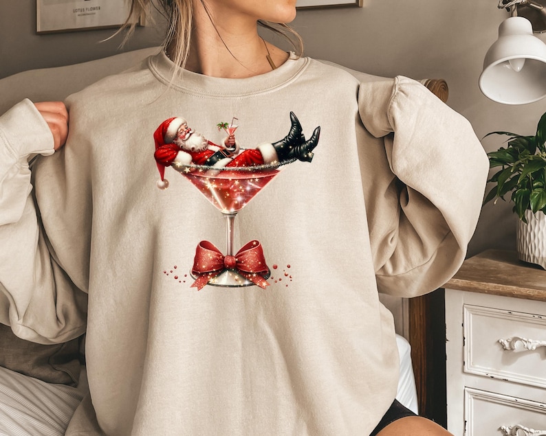 May include: A beige sweatshirt with a graphic of Santa Claus relaxing in a martini glass. Santa is wearing a red suit and a white beard. The glass is filled with a sparkling liquid.