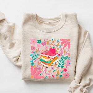 May include: A beige sweatshirt with a colorful graphic print of a stack of books, an apple, and a pencil surrounded by flowers, butterflies, and other whimsical elements.