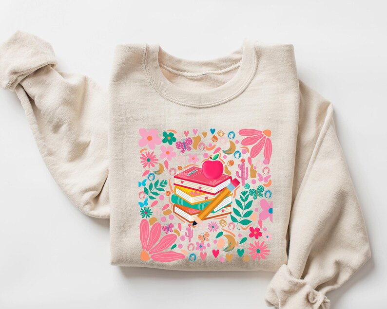 May include: A beige sweatshirt with a colorful graphic print of a stack of books, an apple, and a pencil surrounded by flowers, butterflies, and other whimsical elements.