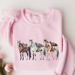 May include: Pink sweatshirt with a graphic of four horses wearing saddles. The horses are brown, white, and black.